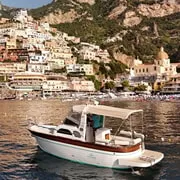 A traditional wooden gozzo departing Praiano marina at dawn for a full-day private boat tour of the Amalfi Coast, with the cliffside village visible above the turquoise sea